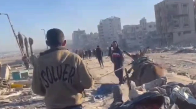 A screenshot from a video showing people walking along a road, with other people lying on a cart being pulled by a donkey