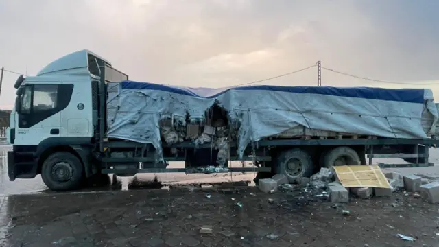 Picture of a lorry, with a canvas trailer. Near the front of the trailer is a large hole where the canvas is torn. Boxes, water bottles and other items are spilling out onto the pavement