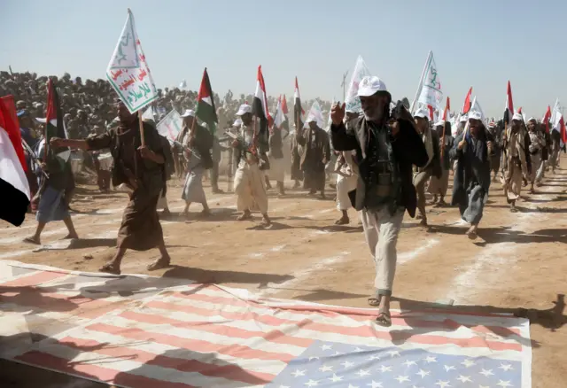 Houthi soldiers and supporters parade in front of a crowd, walking over the flags of the United States and the UK