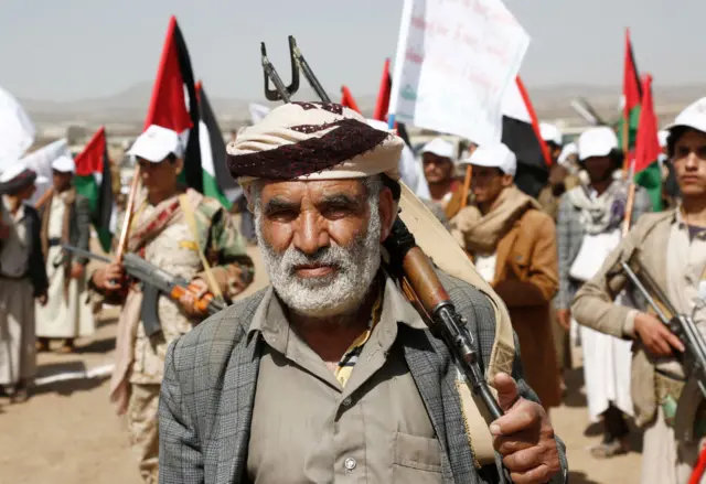 A Houthi fighter carrying a rifle and wearing a traditional turban, with other gun-toting fighters in the background