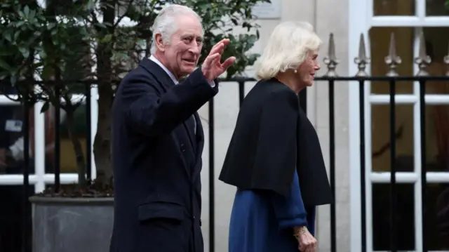 King Charles waves as he and Camilla walk down a street