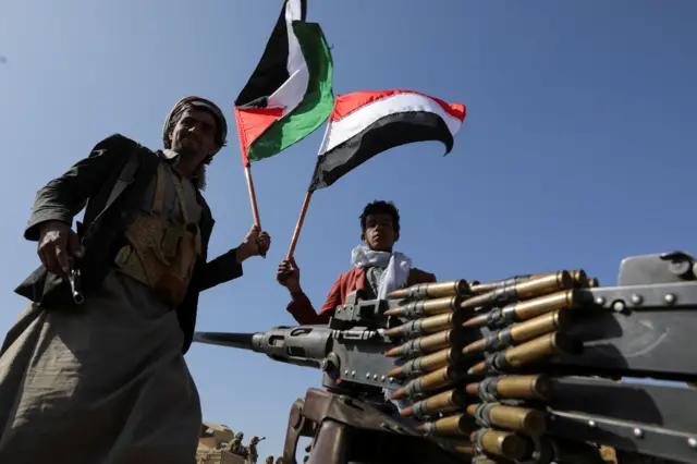 Two Houthi fighters stand around a heavy gun waving the Palestinian and Yemeni flags