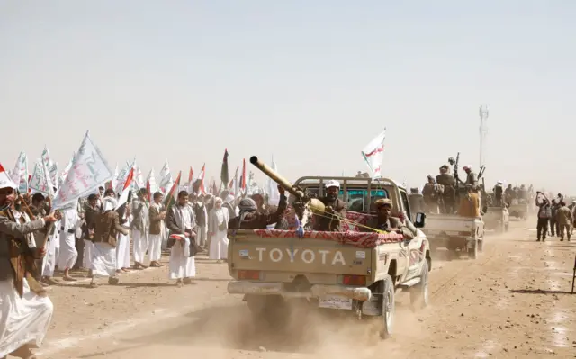 A line of trucks with heavy weapons being driven in front of a flag-waving crowd