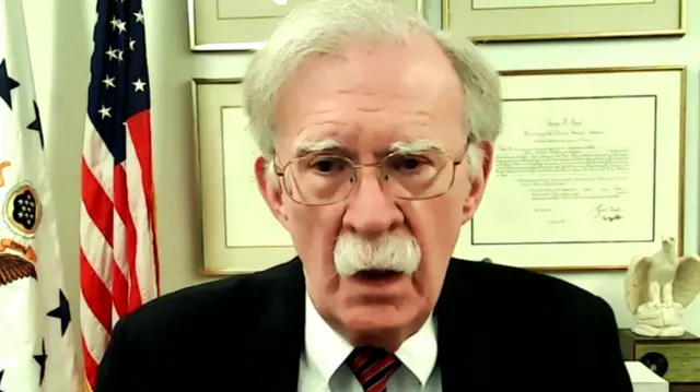 John Bolton speaking on camera. There are frames on the wall and the US flag is behind him.