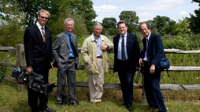 A group of five men with the then-Prince of Wales and Robert Hardman at the centre, with trees in the background