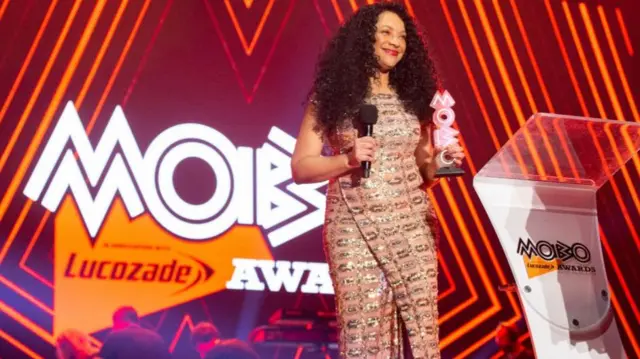A smiling woman with long curly hair stands on a stage with the Mobo logo displayed on a screen behind her