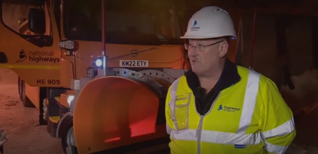 Simon Thain wearing a hard hat speaks in front of a yellow gritter