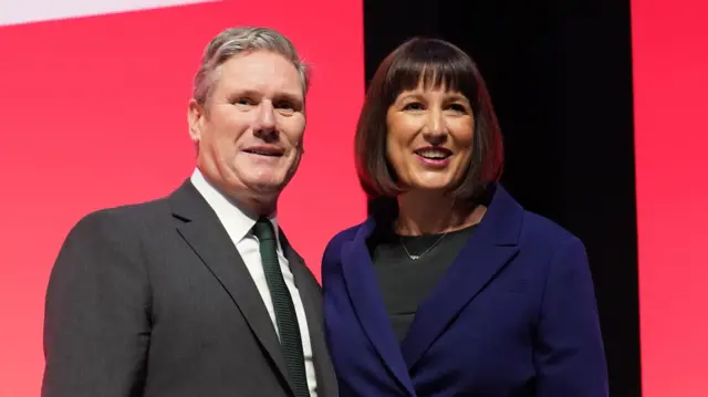 Rachel Reeves and Sir Keir Starmer stand next to each other smiling against a union jack background