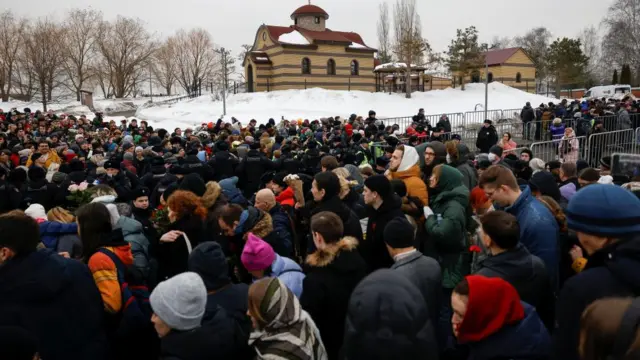 A huge crowd of people in the snow. A chapel sits on a hill nearby