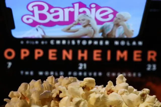 A bucket of popcorn is in the foreground, while the logos of Barbie and Oppenheimer are out of focus behind