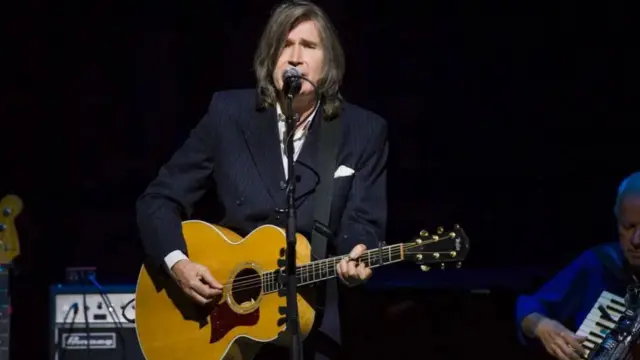 Justin Currie sings into a microphone while playing a guitar as frontman of Del Amitri.