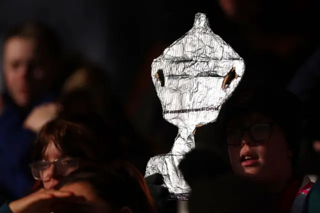 A fan holds up a tinfoil FA Cup in the stands.