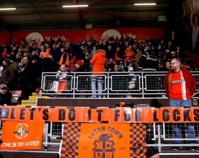 Luton fans stand behind a banner reading 'lets do it for locks' in the away end.