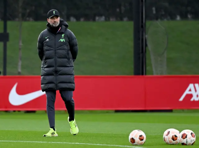 Klopp strolls with his arms behind his back as UEL balls stand on the pitch beside him.