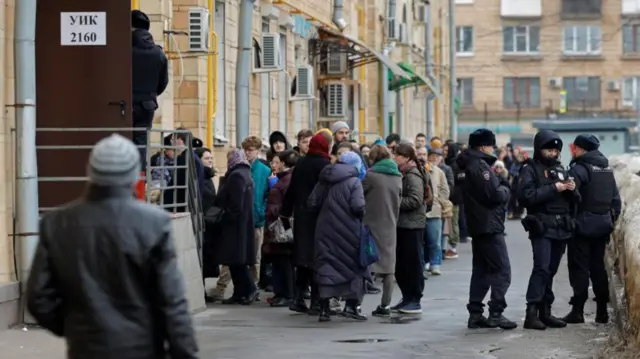 Police officers stand guard next to people, who queue to enter a polling station around noon on the final day of the presidential election in Moscow, Russia, March 17, 2024