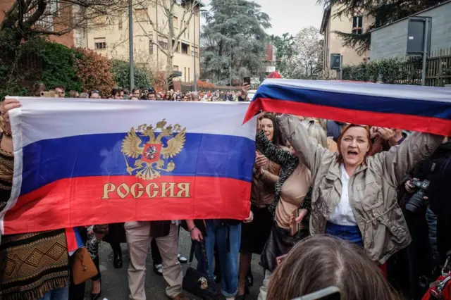 Russians living in Italy hold Russian national flags as they stand in a queue on the last day of voting for the Russian presidential elections, in front of the Russian consulate, in Milan