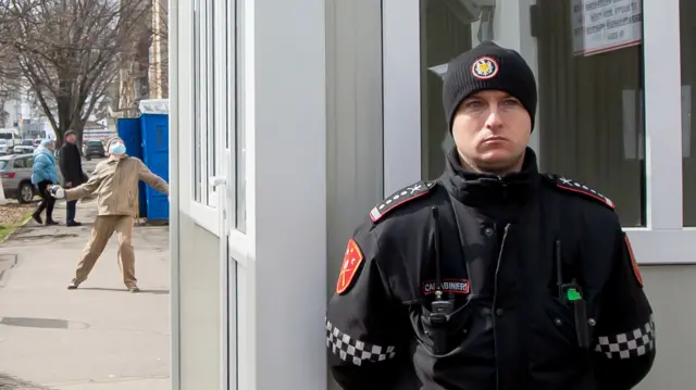 An officer stands guard, unaware that a man wearing a mask in the background is preparing to throw a handmade molotov cocktail towards the Russian Embassy in Chisinau, 17 March 2024