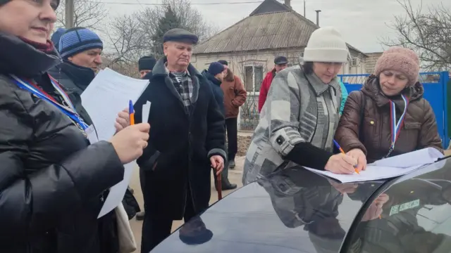 People in Dotesk fill in forms on a car bonnet.