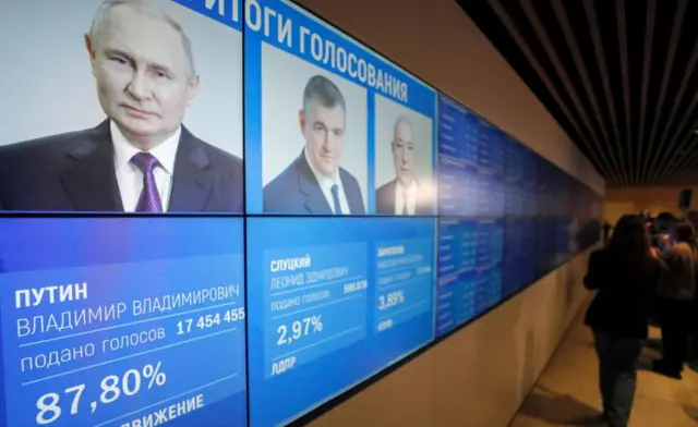 A giant screen shows the presidential candidates. Putin, in the foreground, has 87%