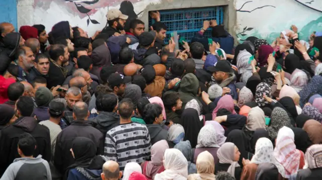 A crowd surrounds a window covered in blue mesh on a concrete building. Those at the front grab the mesh