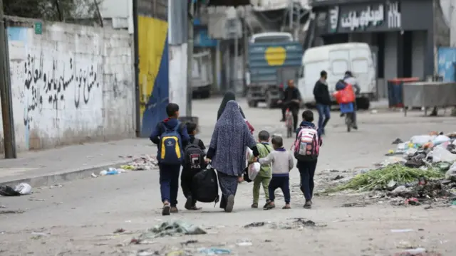 A woman walking with several children, carrying bags with their backs to the camera