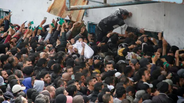 A crowd surges towards a window covered in chicken wire on a concrete building. One man stands above the crowd