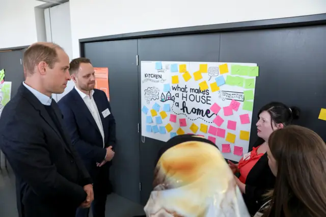 Prince William is shown a poster on which 'what makes a house a home' is written in black font. Post-it notes surround the text