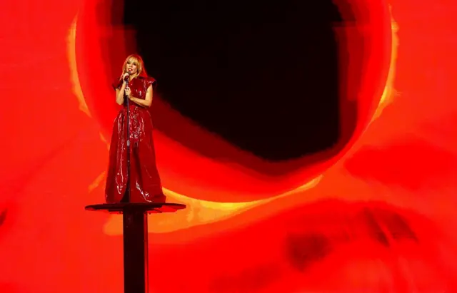 Kylie Minogue performing on a podium with a red backdrop