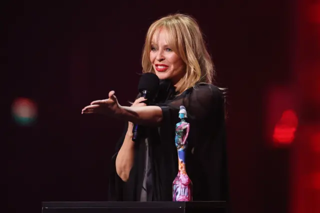 Kylie gestures to her team on stage at the Brits