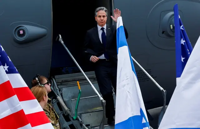 Antony Blinken waving as he gets off the steps of his military plane in Israel, with Israeli and US flags in the foreground