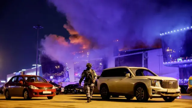 A Russian law enforcement officer walks at a parking area near the burning Crocus City Hall concert venue following a shooting inciden