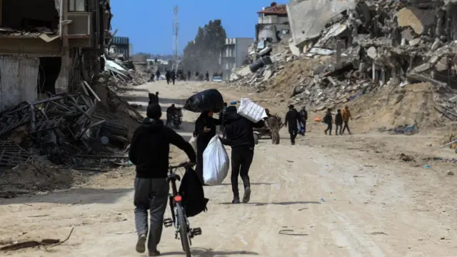 Palestinians with bicycles and other belongs walk down a ruined road with destruction visible on both sides