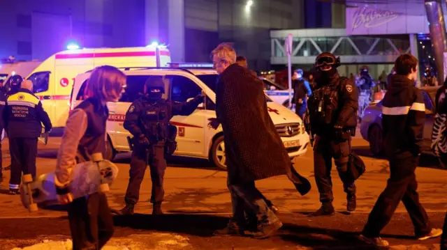 Men cover themselves with blankets while walking past Russian law enforcement officers, who stand guard near the burning Crocus City Hall concert venue following a reported shooting incident, outside Moscow, Russia.