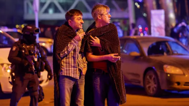 Men cover themselves with blankets as a Russian law enforcement officer stands guard near the burning Crocus City Hall concert venue following the reported shooting incident.