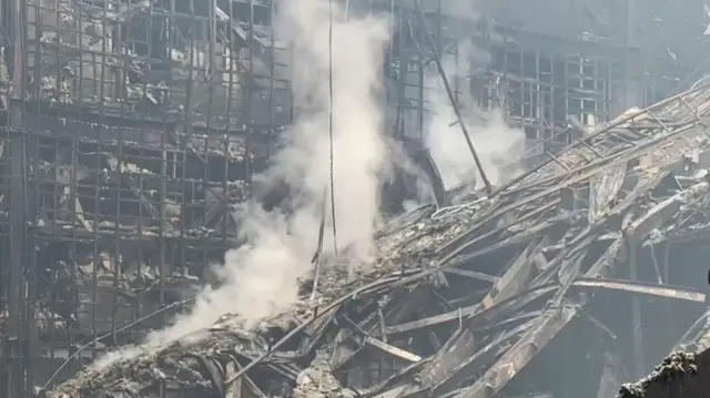 Smoke rises from charred beams and debris.