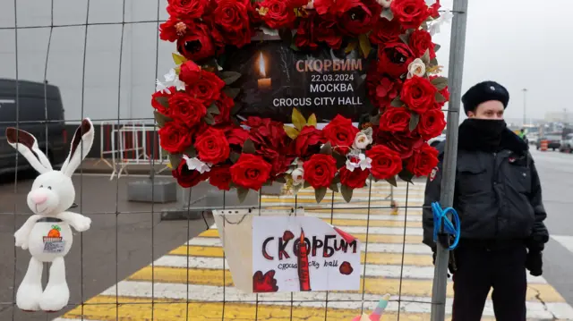 A police officer stabds next to a makeshift memorial on railings made of flowers and white a soft toy rabbit.