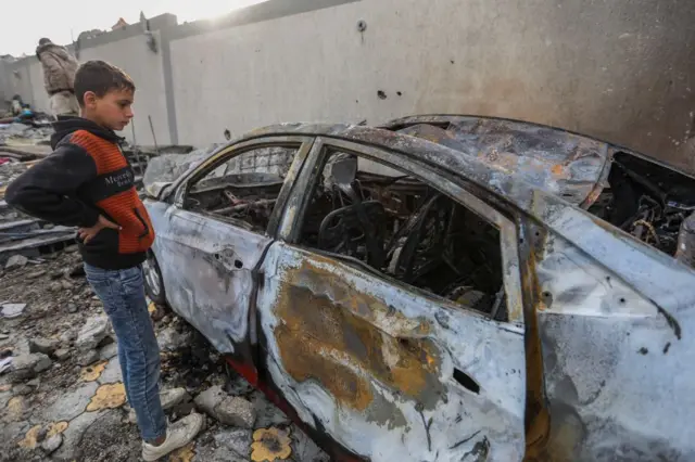 A young boy wearing an orange and black hoodie inspects damage to a car in Rafah