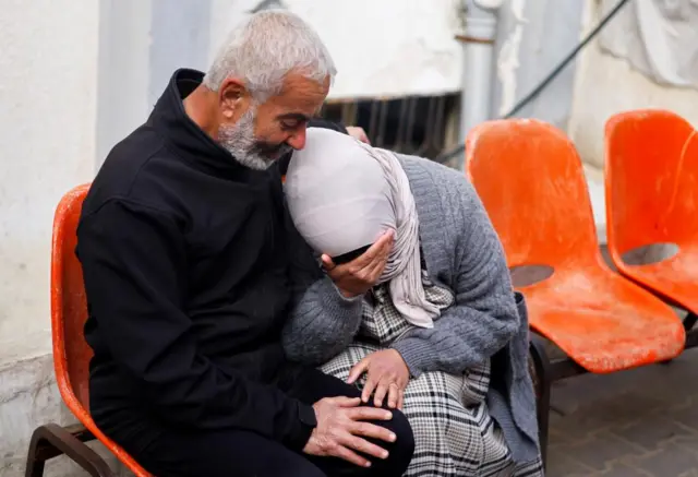 A Palestinian woman wearing a light pink headscarf covers her face with her hand crying as she leans on her husband, who is sitting next to her