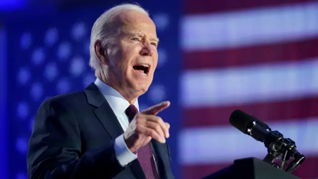 President Joe Biden speaks through a microphone, gestures and stands in front of a projection of a US flag as he holds a campaign rally