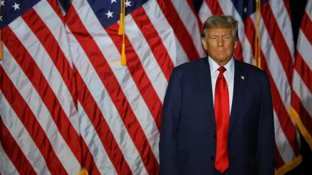 Donald Trump stands in front of a line of US flags, wearing a navy suit and red tie