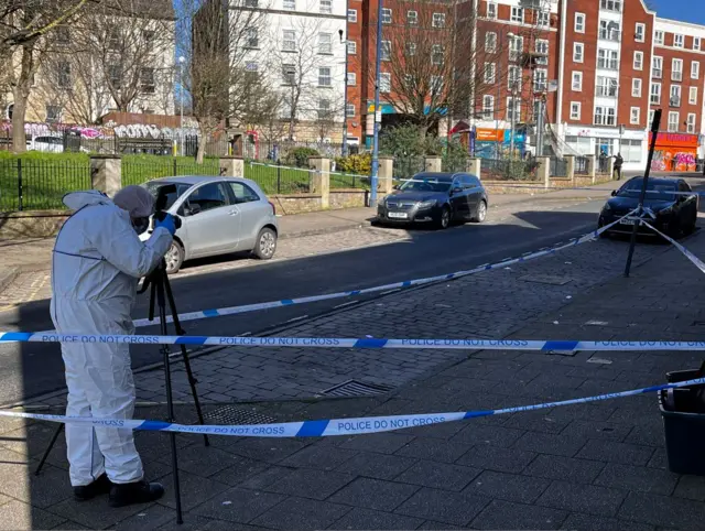 A person in a forensic white suit taking a photograph inside the police cordon