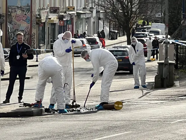 A forensic team in white sanitation suits using tools to dig through drains on the street nearby to the scene