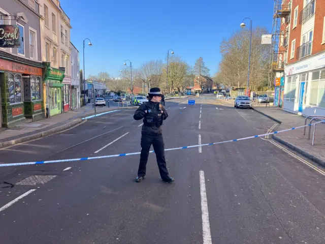 A police woman in her uniform standing in the middle of the road where a police cordon is in place