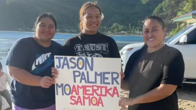 Three Samoan voters stand on a beach and hold a sign that reads "Jason Palmer Mo Amerika Samoa"