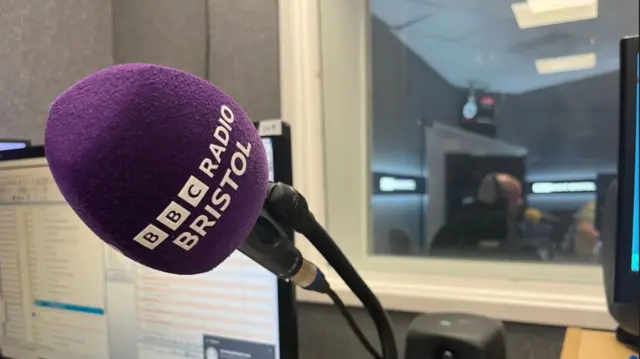 A close up image of the Radio Bristol microphone in the studio, through the window you can see the panel discussing