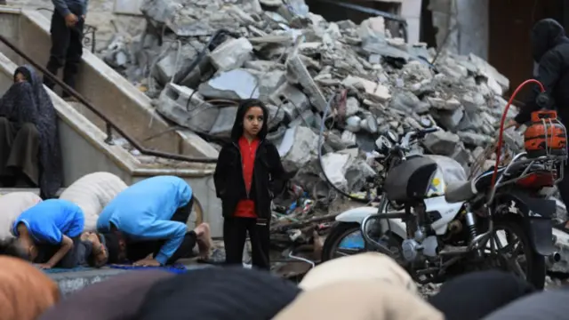 A small Palestianian child standing amidst the ruins of a mosque in Rafah, Gaza whilst muslims pray on the ground next to and in front of them.