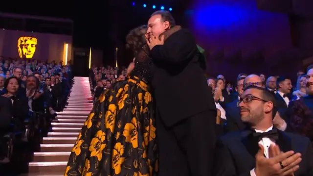 A woman in a yellow flowery dress hugs a man in a suit