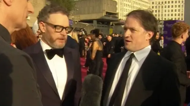 Two men in tuxedos speak to a reporter on a red carpet as the sun sets on the scene