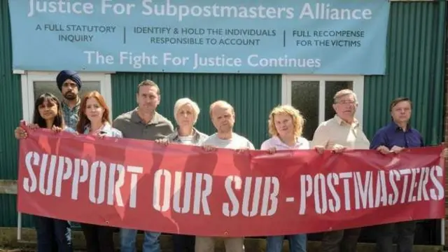 A still from ITV’s Mr Bates vs the Post Officem where all the cast members are lined up and holding a banner which says "support our sub-postmasters"