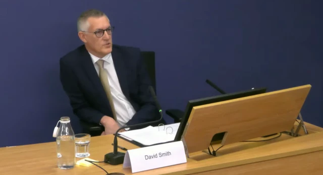 David Smith sat in the inquiry room, leaning to the side at a desk with a sign which reads "David Smith"